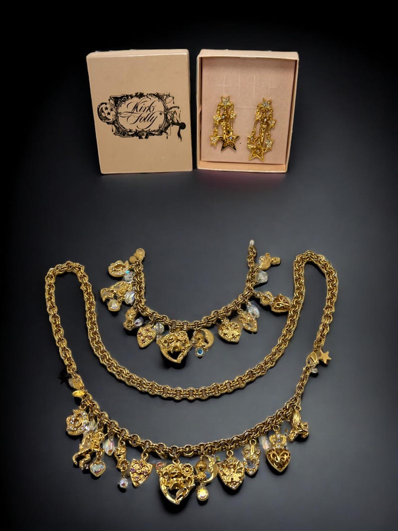 May include: A gold-tone necklace set with multiple charms, including hearts, stars, and other decorative elements. The set also includes a pair of matching earrings. The jewelry is packaged in two pink boxes with the text "Kink & Folly" on one of the boxes.