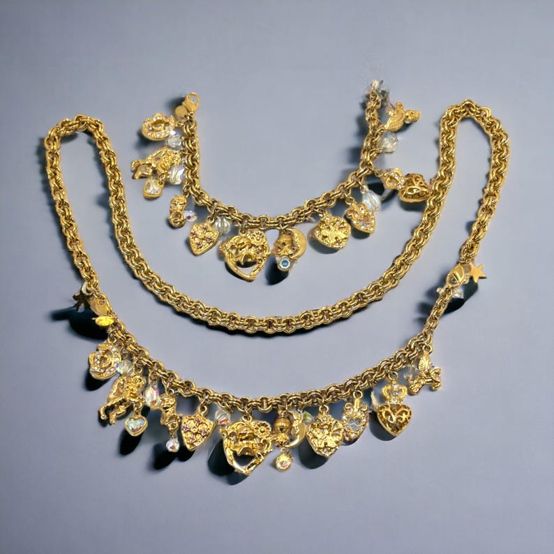 May include: Three gold necklaces with charms, including hearts, stars, and other decorative elements. The necklaces are layered on a gray background.