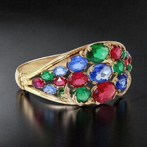 Könnte beinhalten: Ein goldfarbenes Klapparmband mit durchbrochenem Design, das mit zahlreichen oval geschliffenen Edelsteinen in leuchtenden Rot-, Grün- und Blautönen verziert ist, die in einem Clustermuster mit zarten blattartigen Metallakzenten angeordnet sind.