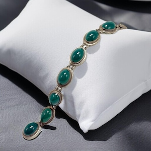 May include: A silver bracelet with oval, emerald-green stones. The stones are set in a silver frame and connected by silver links. The bracelet is displayed on a white pillow.