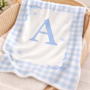 May include: A soft, white baby blanket with a light blue gingham border and a large, light blue letter "A" in the center. The blanket has decorative floral and ribbon accents and is folded in a wicker basket.
