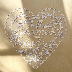 Può includere: Un ritaglio di carta bianco a forma di cuore con un bordo smerlato e dettagli intricati di viticci floreali. Il centro del cuore mostra il testo "home body" in un carattere corsivo, circondato da delicate illustrazioni botaniche.