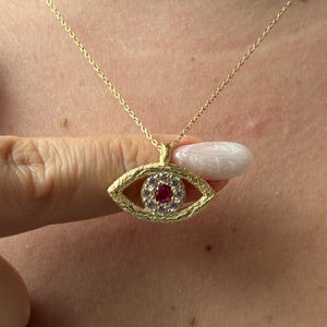 May include: A gold-toned necklace featuring an eye-shaped pendant with a textured finish. The pendant is set with a central round red stone surrounded by a halo of small clear stones, suspended from a delicate gold-toned chain.