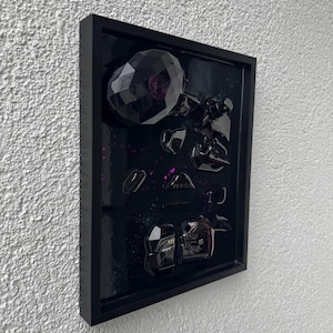 Könnte beinhalten: Ein gerahmtes Kunstwerk mit schwarzem Hintergrund, das schwarze Parfümflaschen und Fragmente zeigt. Das Kunstwerk enthält das Wort "VERSACE" und ist mit violetten Sprenkeln verziert.