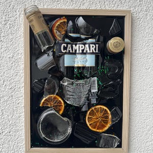 May include: A framed artwork featuring Campari bottles, broken glass, and dried orange slices on a black background. The Campari label is visible, along with bottle fragments and a bottle cap. The frame is a light wood colour.