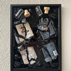 Könnte beinhalten: Ein gerahmtes Kunstwerk mit Fragmenten von Whiskeyflaschen und Etiketten, darunter "Terence Hill" und "Bud Spencer". Die Komposition umfasst klare Glasstücke, Holzstopfen und einen schwarzen Harzhintergrund, wodurch ein einzigartiges Mixed-Media-Stück entsteht.