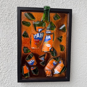 May include: A framed artwork featuring fragments of a green glass bottle and orange Fanta cans. The artwork is set against a textured orange background, with the word "FANTA" visible on the can fragments. The frame is black.