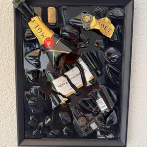 Può includere: Un'opera d'arte da parete incorniciata raffigurante una bottiglia di champagne in vetro verde scuro in frantumi con etichette in lamina d'oro con la scritta Moët & Chandon. I frammenti di vetro sono disposti su uno sfondo nero lucido con un tappo.