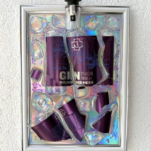 May include: A framed wall art piece featuring a Rammstein Gin bottle with a purple label, 27% alc/vol, 700 ml, embedded in a holographic, iridescent resin background with scattered purple glitter and clear, jagged, glass-like shards.