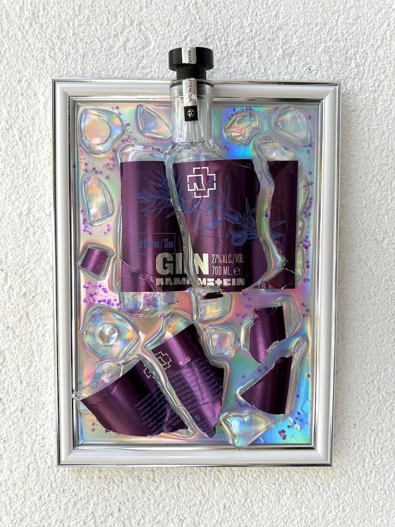 May include: A framed wall art piece featuring a Rammstein Gin bottle with a purple label, 27% alc/vol, 700 ml, embedded in a holographic, iridescent resin background with scattered purple glitter and clear, jagged, glass-like shards.
