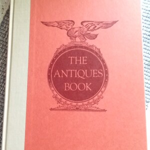 May include: The cover of a red book with a gold spine. The book title is "The Antiques Book" and features a stylized eagle with a shield.