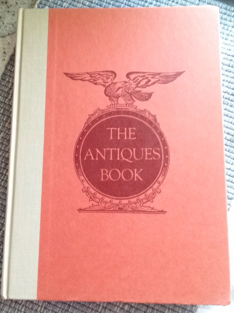 May include: The cover of a red book with a gold spine. The book title is "The Antiques Book" and features a stylized eagle with a shield.