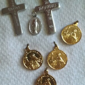 May include: Collection of religious pendants and crosses. Includes two silver crosses with engraved text, a silver oval pendant with a figure, and several gold-colored round pendants with a saintly figure.
