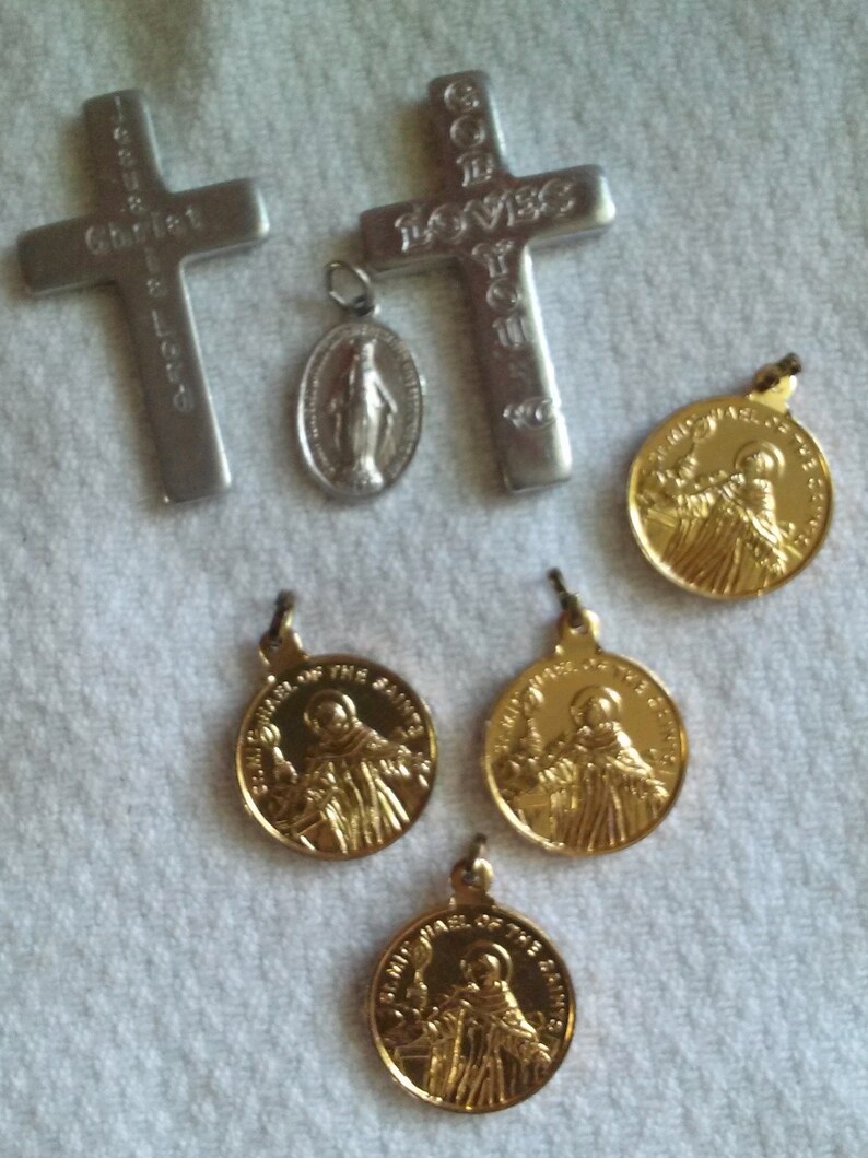 May include: Collection of religious pendants and crosses. Includes two silver crosses with engraved text, a silver oval pendant with a figure, and several gold-colored round pendants with a saintly figure.