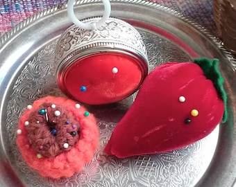 May include: Three pincushions on a silver tray. One is a round, red pincushion with a silver lid and a white ring handle. Another is a red, velvet strawberry-shaped pincushion. The third is a crocheted pincushion with a brown center and orange outer layer.