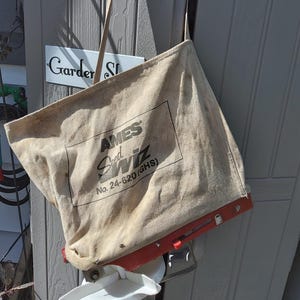 May include: A beige canvas seed spreader bag featuring the black printed text AMES Seed Wiz No. 24-620 (GHS). The bag is attached to a red plastic base with an adjustable orange lever and a white plastic dispensing component at the bottom.