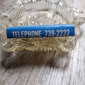 May include: A blue and white rectangular sticker with the text "TELEPHONE 739-2222" on a clear glass ashtray.
