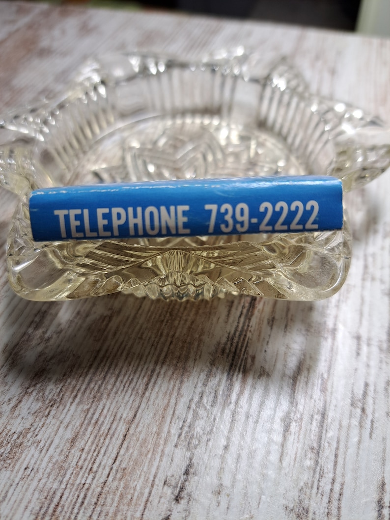 May include: A blue and white rectangular sticker with the text "TELEPHONE 739-2222" on a clear glass ashtray.