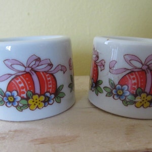 May include: Two white ceramic egg cups with a colorful Easter egg design. The eggs are red, orange, yellow, and pink with a white ribbon and green leaves. There are also blue and yellow flowers.