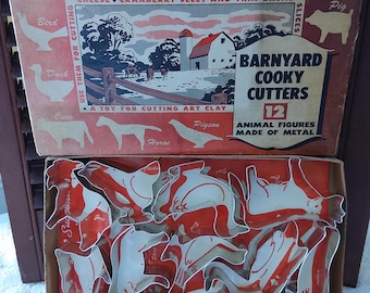 以下が含まれることがあります： 赤と白の箱に「Barnyard Cooky Cutters 12 Animal Figures Made of Metal」と書いてあります。箱が開いていて、農場の動物の形をした金属製のクッキーカッターが12個入っています。
