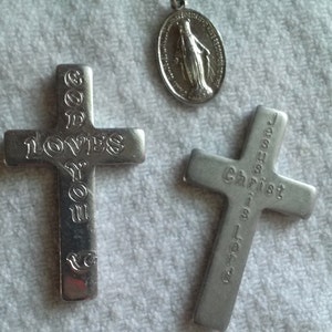 May include: Three silver religious pendants. Two crosses with engraved text: "GOD LOVES YOU" and "JESUS Christ is Lord". A circular pendant depicts a religious figure. All pendants are on a white, textured surface.