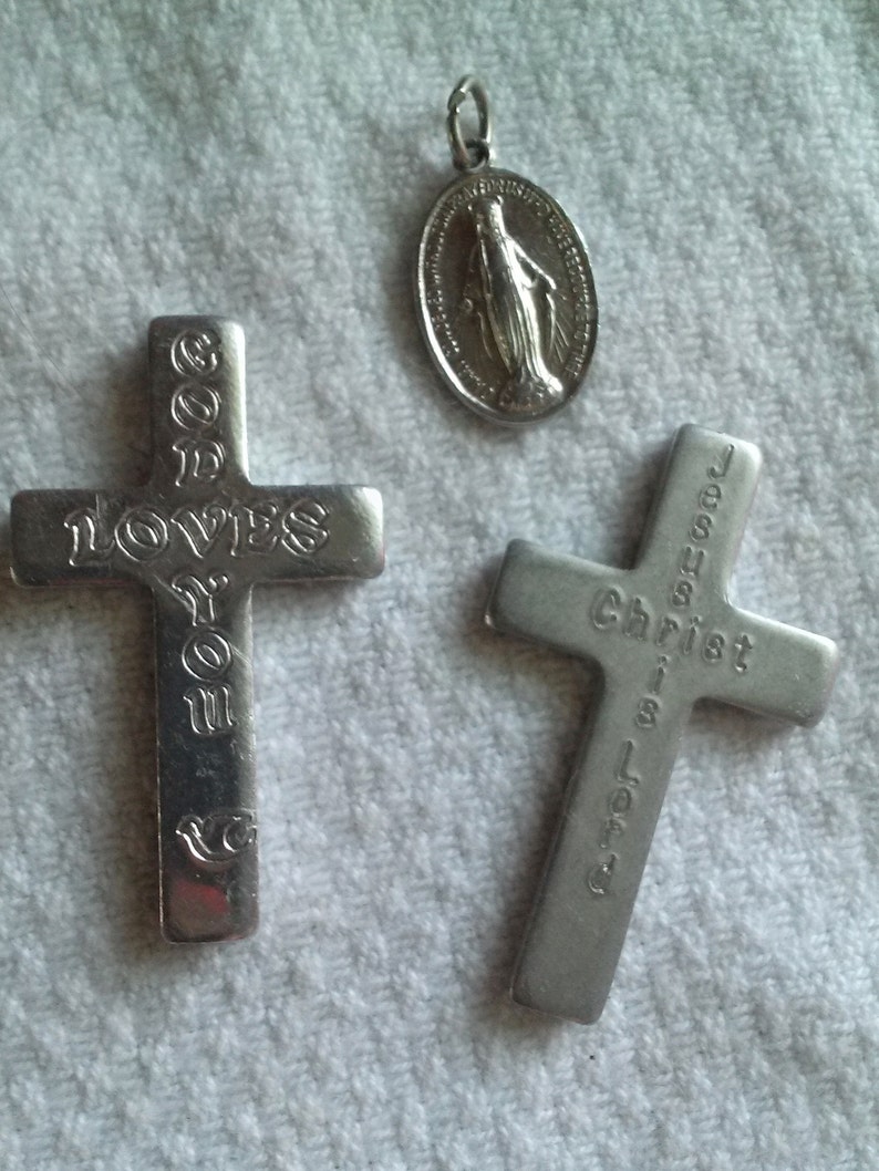 May include: Three silver religious pendants. Two crosses with engraved text: "GOD LOVES YOU" and "JESUS Christ is Lord". A circular pendant depicts a religious figure. All pendants are on a white, textured surface.