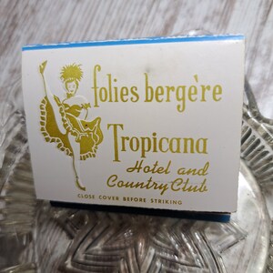 May include: A white matchbook with a blue border. The matchbook features a gold illustration of a woman in a dance costume and the text "folies berg&egrave;re Tropicana Hotel and Country Club". The text "Close cover before striking" is printed at the bottom.
