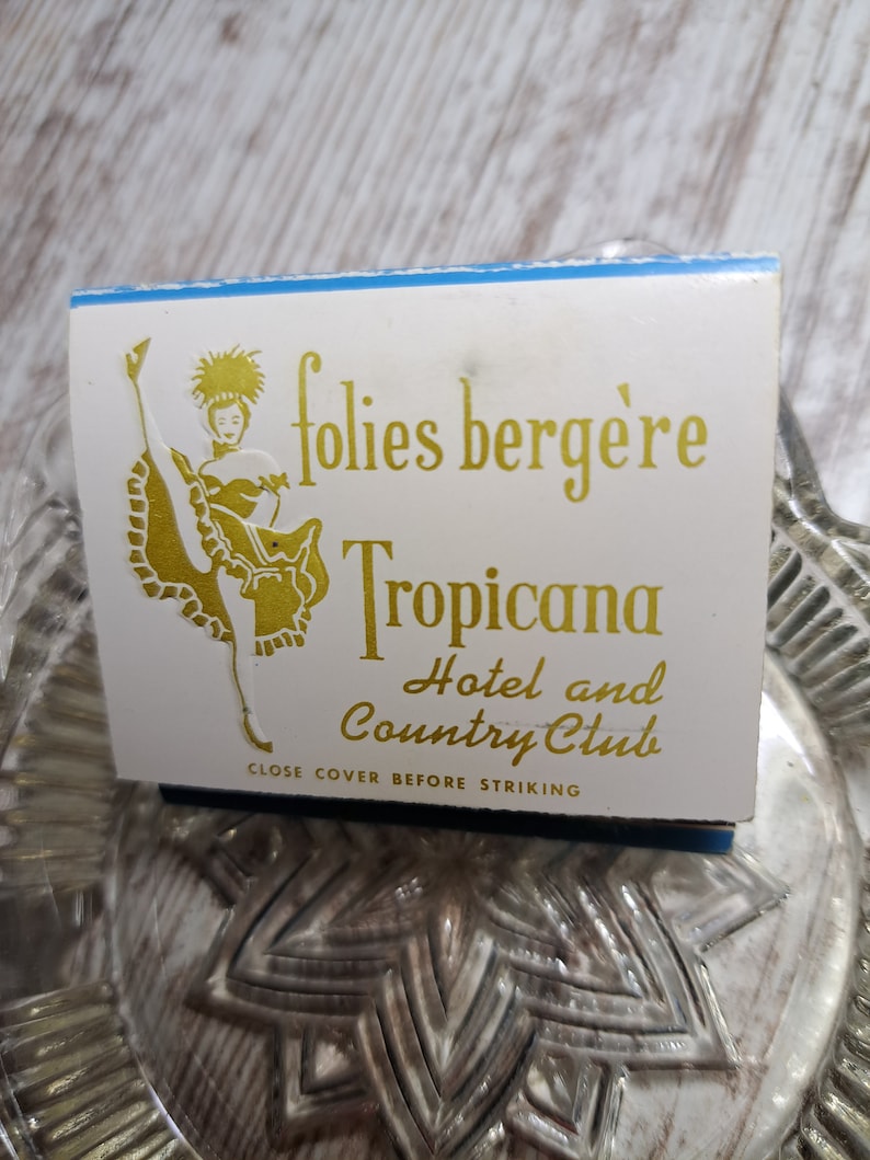May include: A white matchbook with a blue border. The matchbook features a gold illustration of a woman in a dance costume and the text "folies berg&egrave;re Tropicana Hotel and Country Club". The text "Close cover before striking" is printed at the bottom.