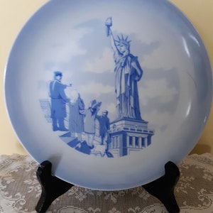 May include: A blue and white ceramic plate with a depiction of the Statue of Liberty and four figures standing in front of it. The plate is on a white lace doily and a black stand.