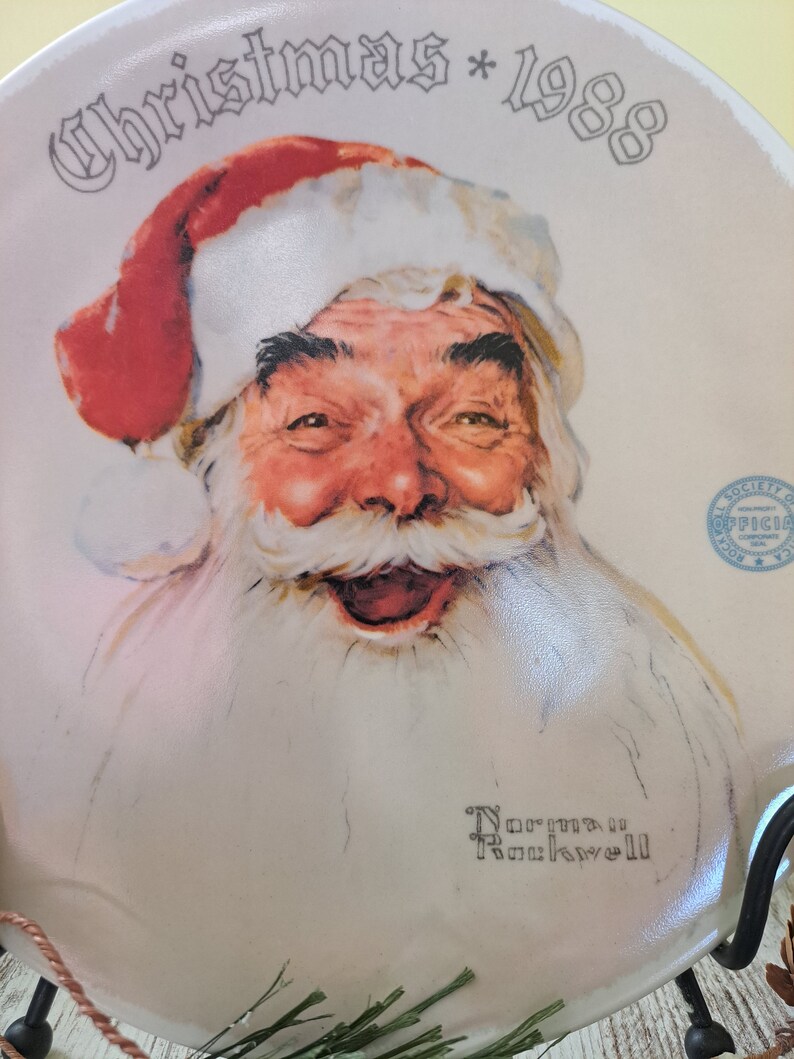 May include: A white ceramic plate with a vintage illustration of Santa Claus. The plate has the text "Christmas * 1988" and "Norman Rockwell" in black lettering.
