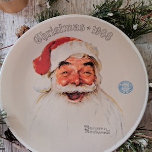 May include: A white ceramic plate with a vintage illustration of Santa Claus. The plate has the text "Christmas * 1988" and "Norman Rockwell" printed on it.