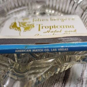 May include: A blue and white matchbook with gold lettering. The matchbook advertises the Tropicana Hotel and Country Club in Las Vegas, Nevada. The matchbook is made by the American Match Co.