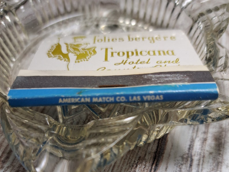 May include: A blue and white matchbook with gold lettering. The matchbook advertises the Tropicana Hotel and Country Club in Las Vegas, Nevada. The matchbook is made by the American Match Co.