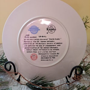 May include: A white ceramic plate with a blue and red border. The plate features a 1988 limited edition print of "Santa Claus" by Norman Rockwell. The plate is numbered 9041C and is certified by the Rockwell Society of America. The plate is permanently closed by Edwin M. Knowles on December 31, 1988.