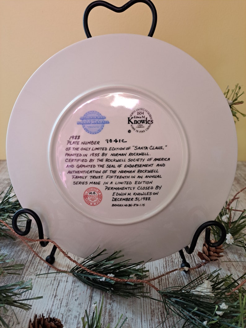 May include: A white ceramic plate with a blue and red border. The plate features a 1988 limited edition print of "Santa Claus" by Norman Rockwell. The plate is numbered 9041C and is certified by the Rockwell Society of America. The plate is permanently closed by Edwin M. Knowles on December 31, 1988.