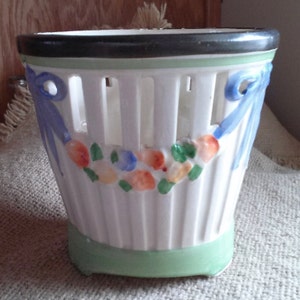 May include: White ceramic planter with a green base and blue ribbon accents. The planter has a lattice design and is decorated with painted flowers.