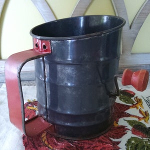 May include: A vintage metal flour sifter with a red handle and a red wooden crank. The sifter has a gray finish and is sitting on a fabric surface with a rooster design.