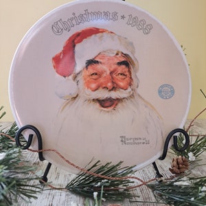 May include: A white ceramic plate with a festive Christmas design featuring a smiling Santa Claus. The plate has the text "Christmas * 1988" printed on it. The plate is displayed on a black metal stand.