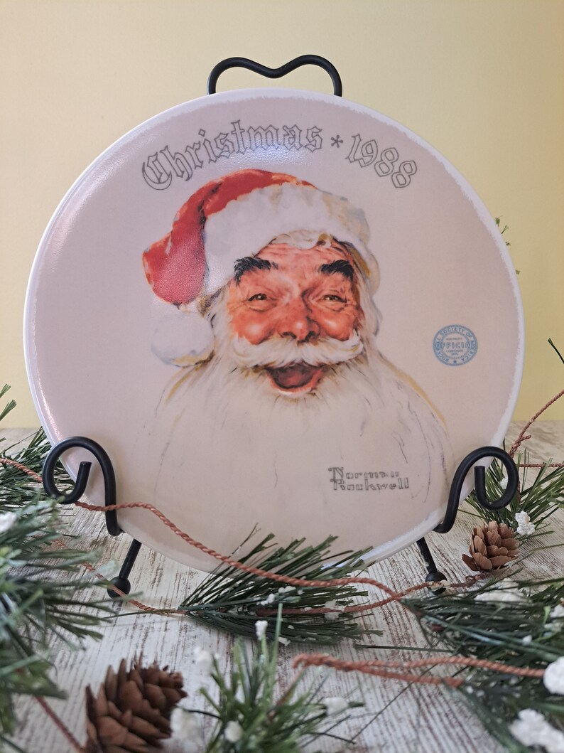May include: A white ceramic plate with a festive Christmas design featuring a smiling Santa Claus. The plate has the text "Christmas * 1988" printed on it. The plate is displayed on a black metal stand.