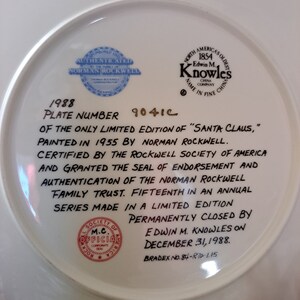 May include: A white ceramic plate with a blue and gold border. The plate features a black and white illustration of Santa Claus. The back of the plate has text that reads "1988 Plate Number 9041C of the only limited edition of "Santa Claus," painted in 1955 by Norman Rockwell. Certified by the Rockwell Society of America and granted the seal of endorsement and authentication of the Norman Rockwell Family Trust. Fifteenth in an annual series made in a limited edition permanently closed by Edwin M. Knowles on December 31, 1988. Bradex No. 84-870-1.15"