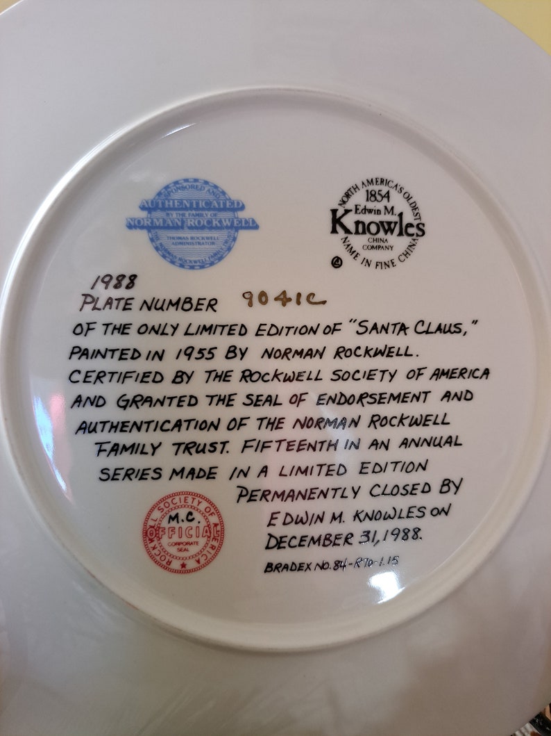 May include: A white ceramic plate with a blue and gold border. The plate features a black and white illustration of Santa Claus. The back of the plate has text that reads "1988 Plate Number 9041C of the only limited edition of "Santa Claus," painted in 1955 by Norman Rockwell. Certified by the Rockwell Society of America and granted the seal of endorsement and authentication of the Norman Rockwell Family Trust. Fifteenth in an annual series made in a limited edition permanently closed by Edwin M. Knowles on December 31, 1988. Bradex No. 84-870-1.15"