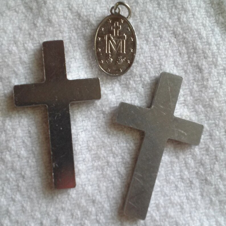 May include: Three silver religious pendants. Two are crosses, and the third is an oval pendant with the letter "M" and other symbols. The pendants are displayed on a white, textured surface.