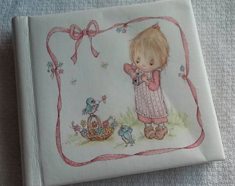 May include: A white photo album with a pink ribbon border. The album features a drawing of a girl with blonde hair holding a camera, and two blue birds in a basket with flowers.