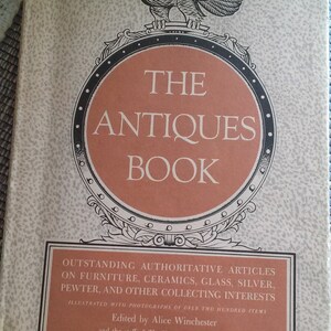 May include: The cover of a book titled "The Antiques Book" with an illustration of an eagle perched on a decorative crest. The book features articles on furniture, ceramics, glass, silver, pewter, and other collecting interests. It is illustrated with photographs of over two hundred items and edited by Alice Winchester and the staff of The Magazine Antiques.