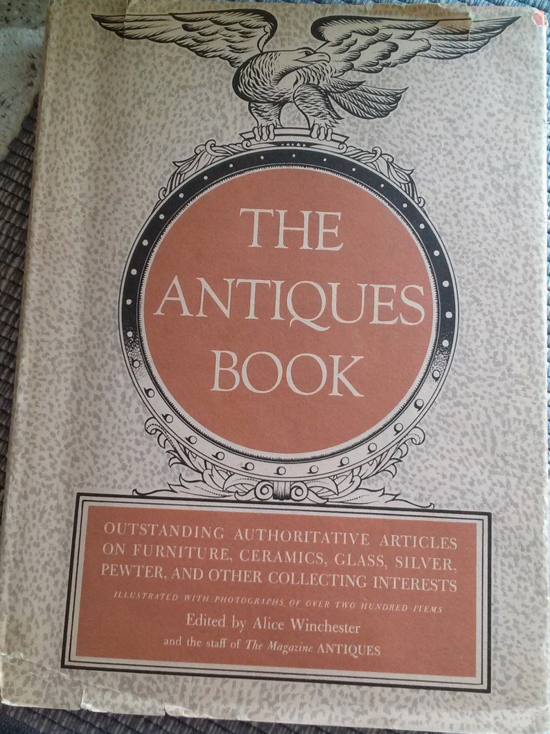 May include: The cover of a book titled "The Antiques Book" with an illustration of an eagle perched on a decorative crest. The book features articles on furniture, ceramics, glass, silver, pewter, and other collecting interests. It is illustrated with photographs of over two hundred items and edited by Alice Winchester and the staff of The Magazine Antiques.