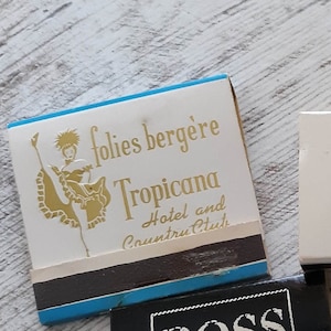 May include: A vintage matchbook cover with a blue and white design. It features a gold illustration of a woman in a dance pose and the text "folies berg&egrave;re Tropicana Hotel and Country Club".