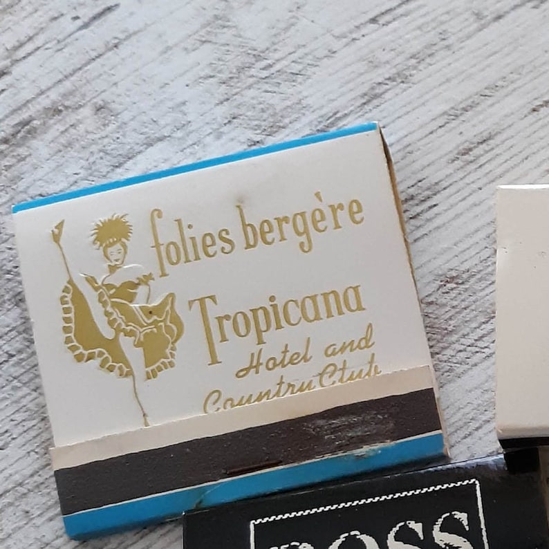 May include: A vintage matchbook cover with a blue and white design. It features a gold illustration of a woman in a dance pose and the text "folies berg&egrave;re Tropicana Hotel and Country Club".