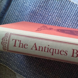 May include: A red and cream colored book titled "The Antiques Book" by Alice Winchester.