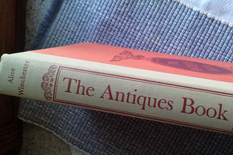 May include: A red and cream colored book titled "The Antiques Book" by Alice Winchester.