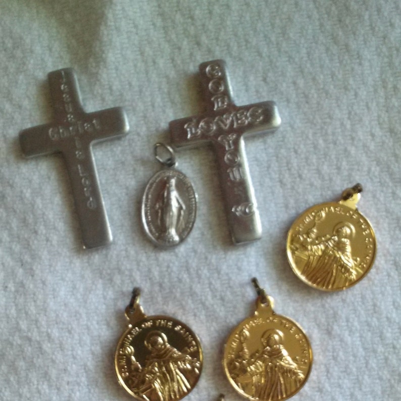May include: An assortment of religious pendants and crosses. Two silver-colored crosses with engraved text, one reading "Christ" and the other "God Loves You." A small oval pendant with a religious figure and several gold-colored medallions.